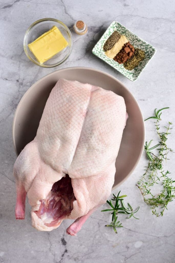 Whole raw duck with herbs, spices, and butter on a marble surface, ready for cooking preparation.
