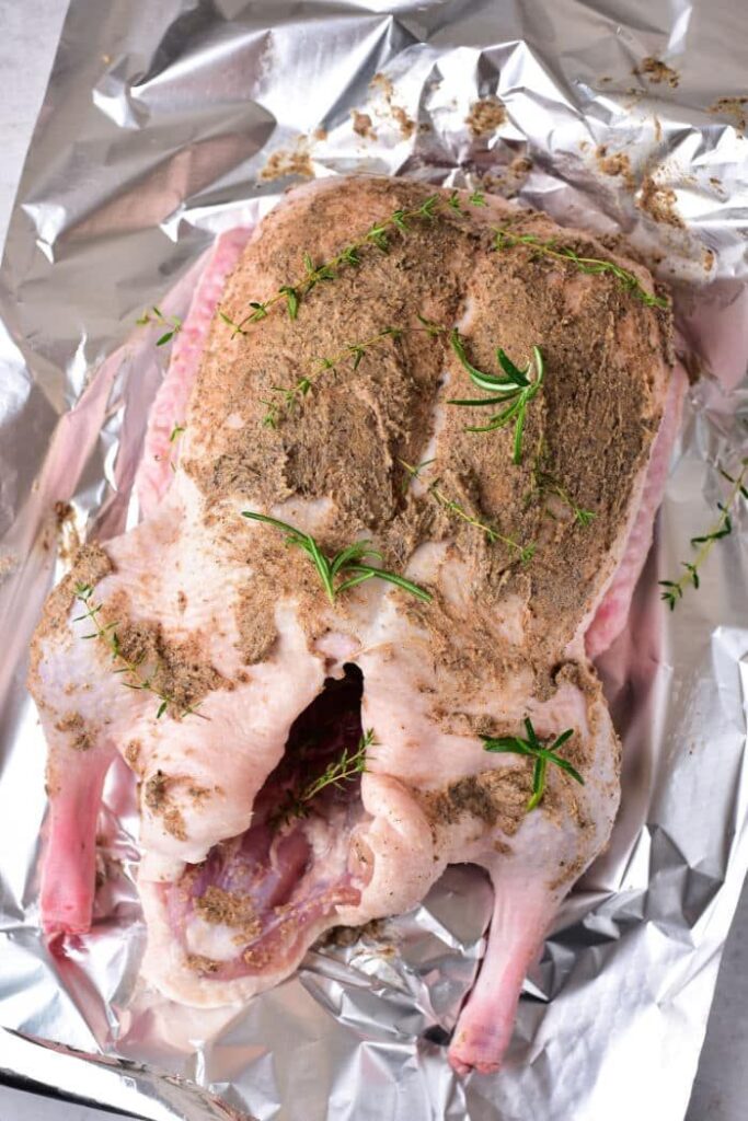 Raw seasoned duck on aluminum foil ready for oven roasting with herbs and spices.