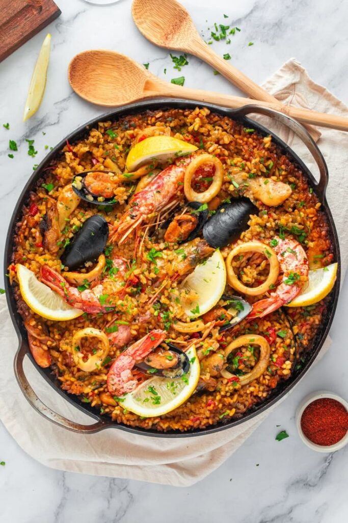 Traditional seafood paella with shrimp, mussels, and lemon slices in a pan on a marble table.