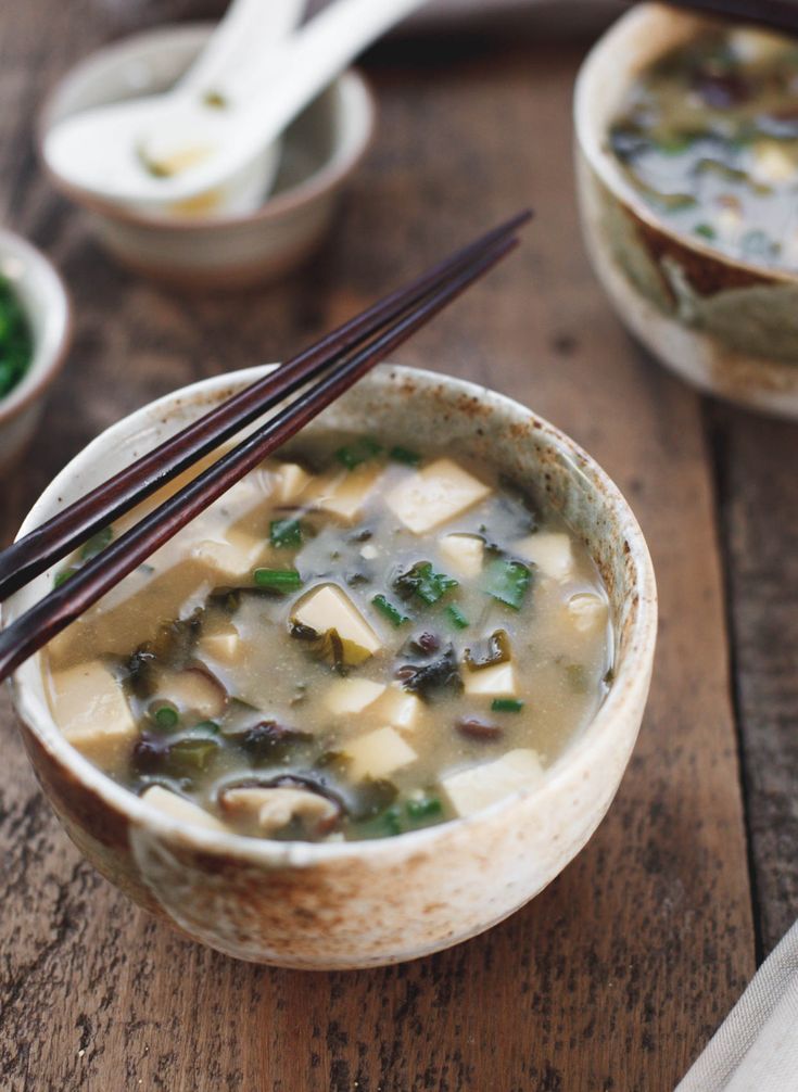 Bowl of tofu miso soup with seaweed and green onions on a wooden table, with chopsticks resting on the edge.