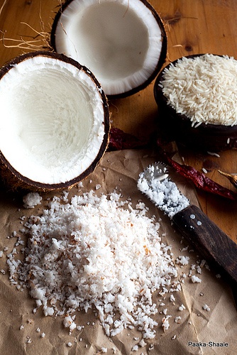 Shredded coconut with open coconut shells, rice, and a knife on a wooden table.