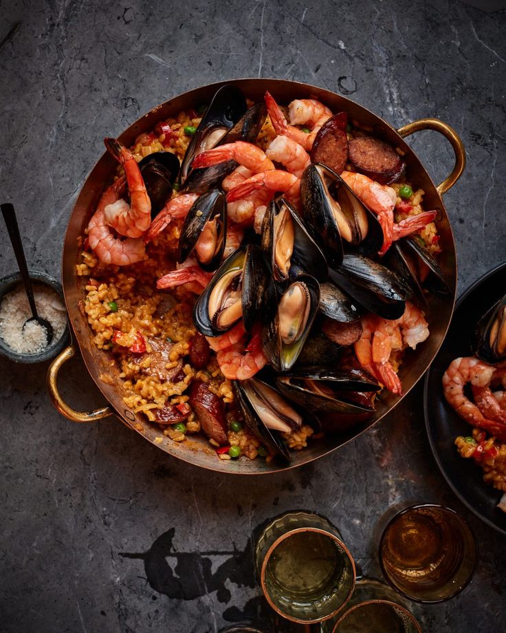 Seafood paella with shrimp, mussels, and chorizo in a pan on a dark table, accompanied by salt and a glass of drink.