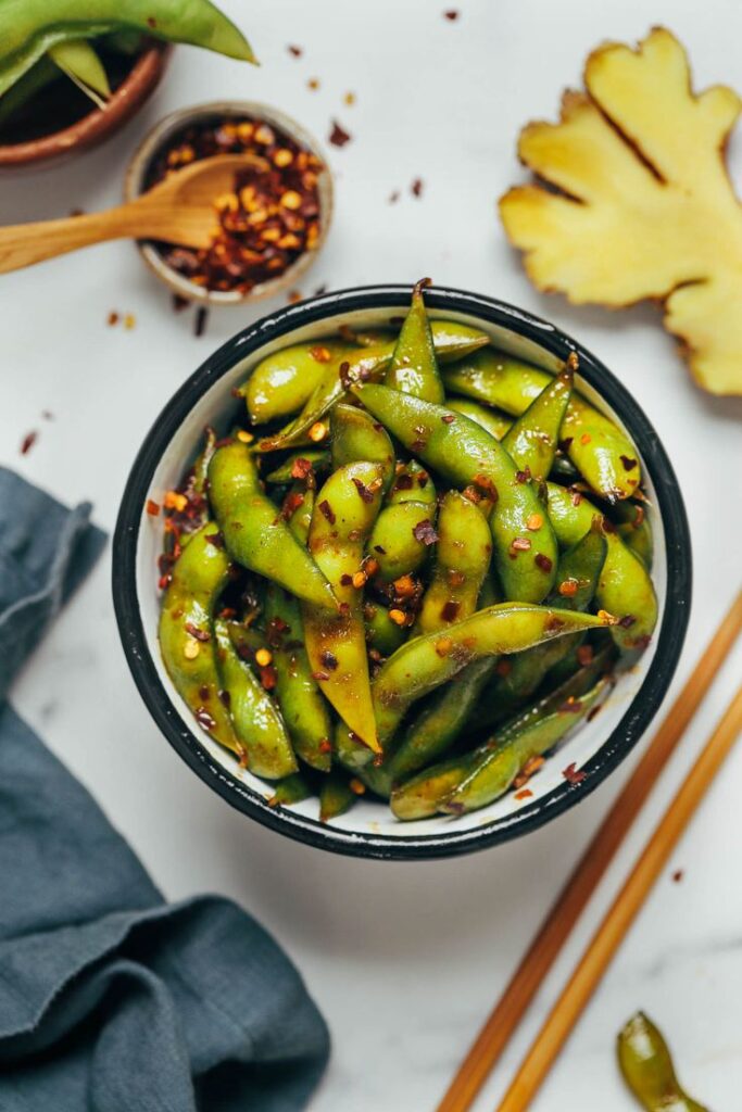 Spicy edamame in a bowl with chili flakes, ginger, and chopsticks on a marble table. Asian-inspired snack.