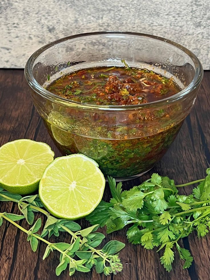 Glass bowl of salsa verde with lime halves and fresh herbs on a wooden table.