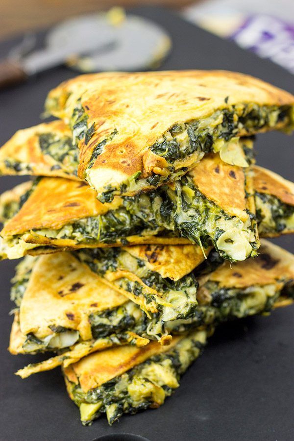 Stacked spinach and cheese quesadillas on a black surface, ready to serve with a pizza cutter in the background.