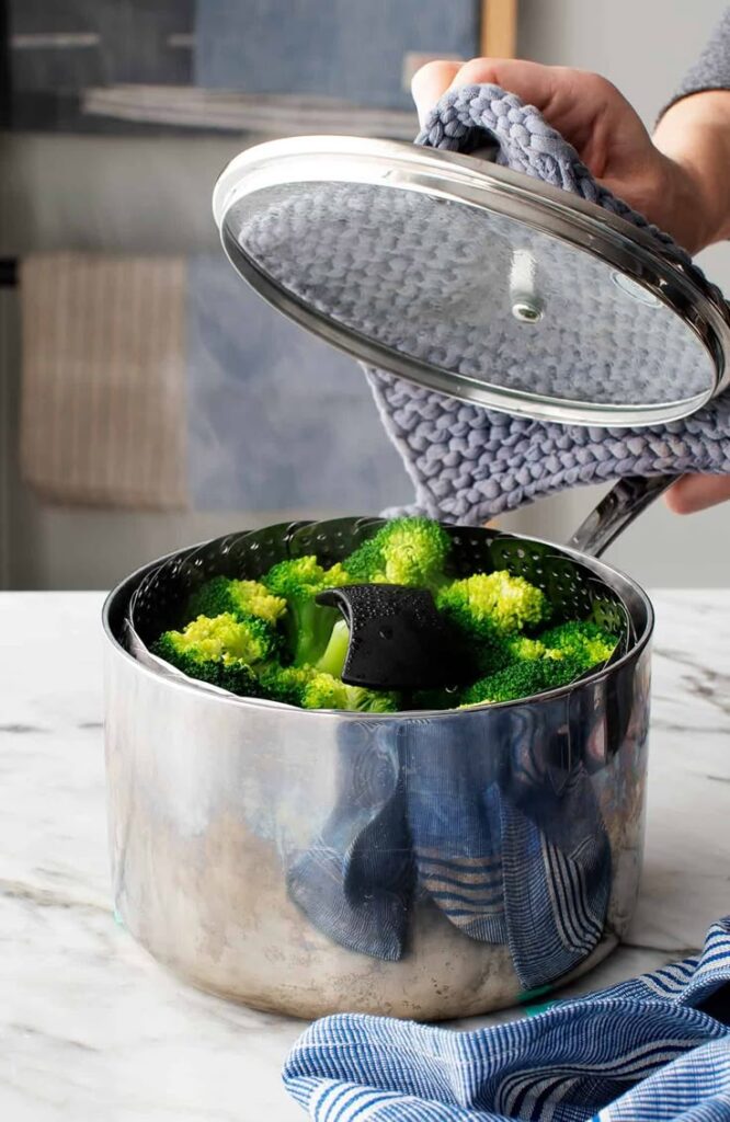 Steamed broccoli in a pot with lid being lifted by hand with oven mitt. Healthy cooking concept.