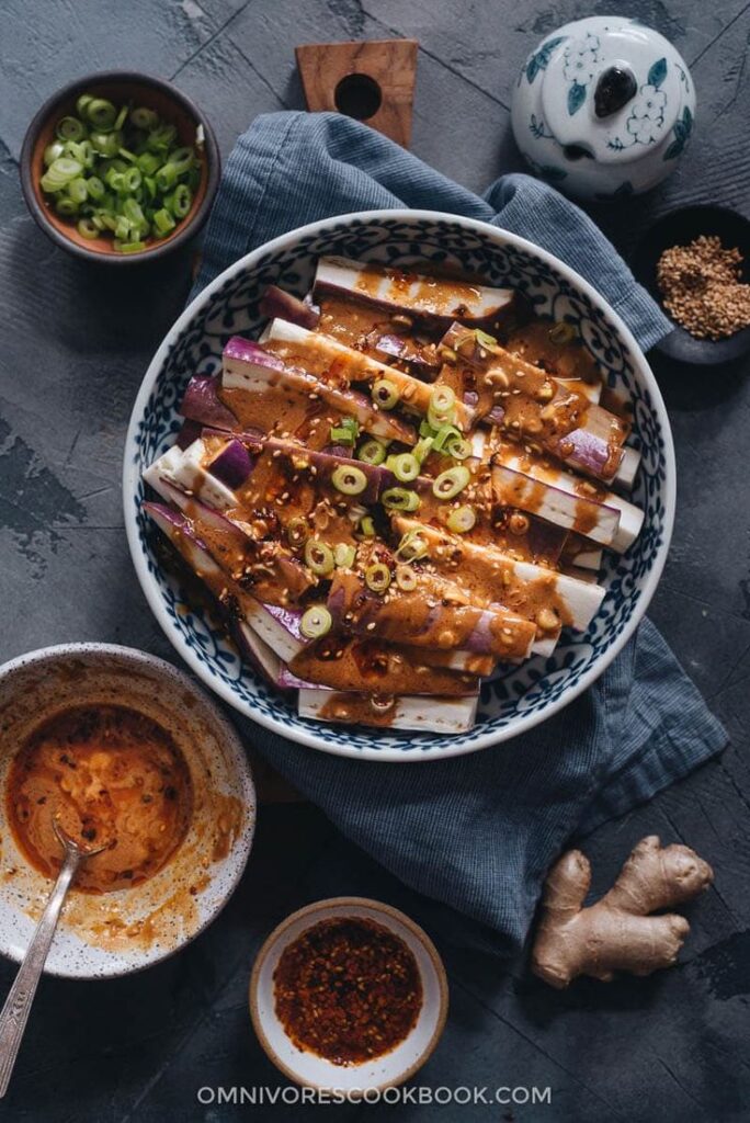 Sliced eggplant with spicy peanut sauce and green onions in a bowl; ingredients and sauces surrounding the dish.