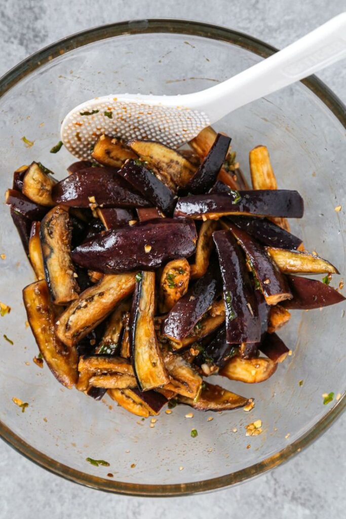 Stir-fried eggplant slices in a glass bowl with a white spatula, seasoned with herbs and spices.