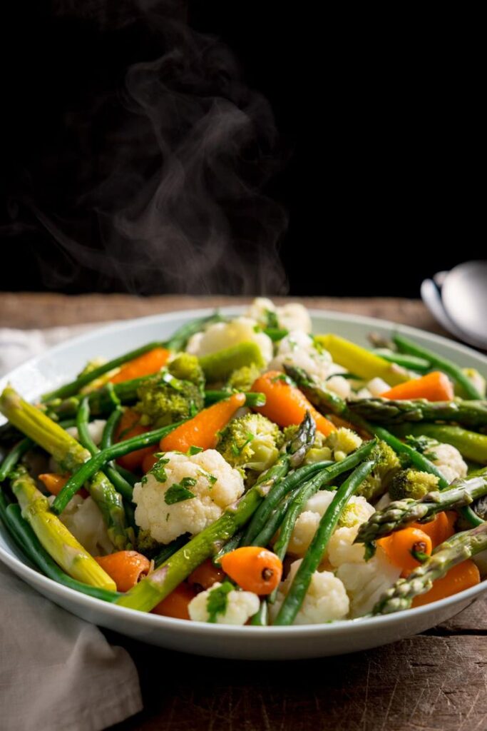 Steaming plate of mixed steamed vegetables including asparagus, carrots, cauliflower, and broccoli on a wooden table.
