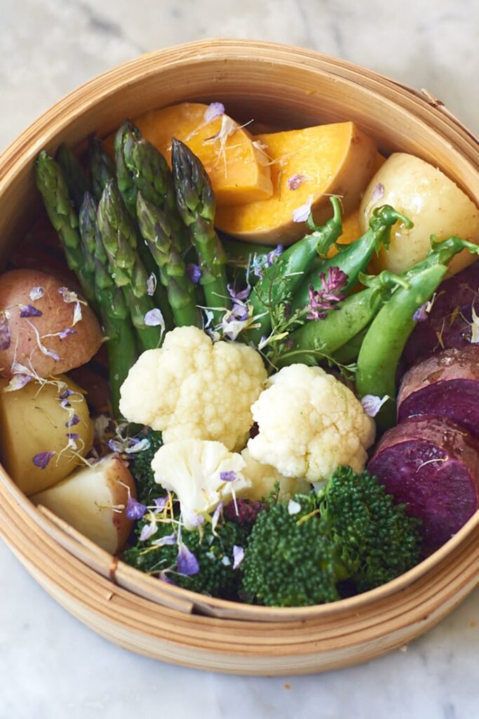Fresh steamed vegetables in a bamboo basket: asparagus, cauliflower, broccoli, potatoes, and squash with flowers.