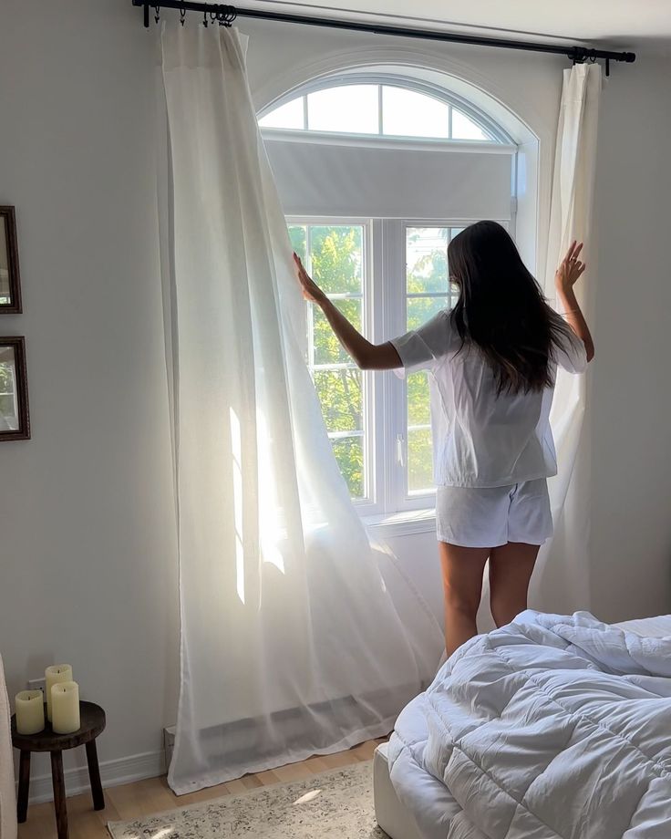 Woman in white pajamas opening curtains to let morning light into a cozy bedroom with candles and a soft bed.