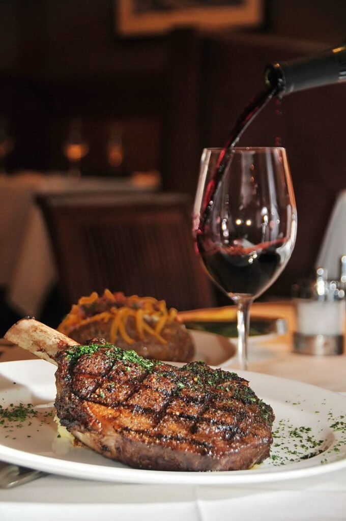 Grilled ribeye steak on a plate with a glass of red wine being poured in a cozy restaurant setting.