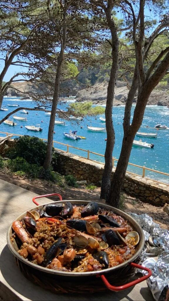 Seafood paella by the seaside with boats and scenic coastal view through trees at a picturesque outdoor setting.