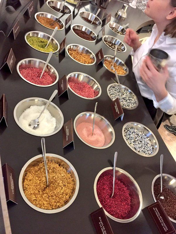 Assorted toppings for desserts in metal bowls with spoons at a counter with a staff member assisting.