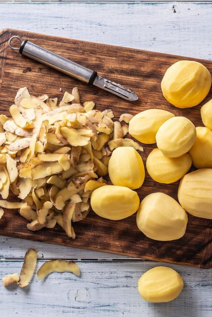 Peeled potatoes and potato peels on a wooden cutting board with a peeler, ready for cooking.