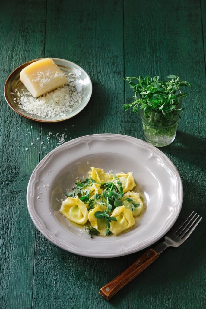 Plate of tortellini with fresh herbs and grated cheese on green table, next to a block of cheese and a herb jar.