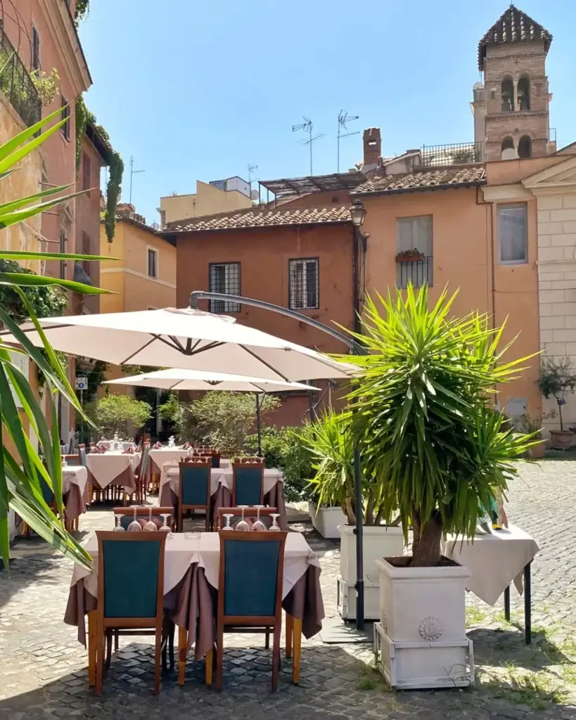 Charming outdoor cafe in European courtyard with rustic buildings and greenery under sunny blue sky.