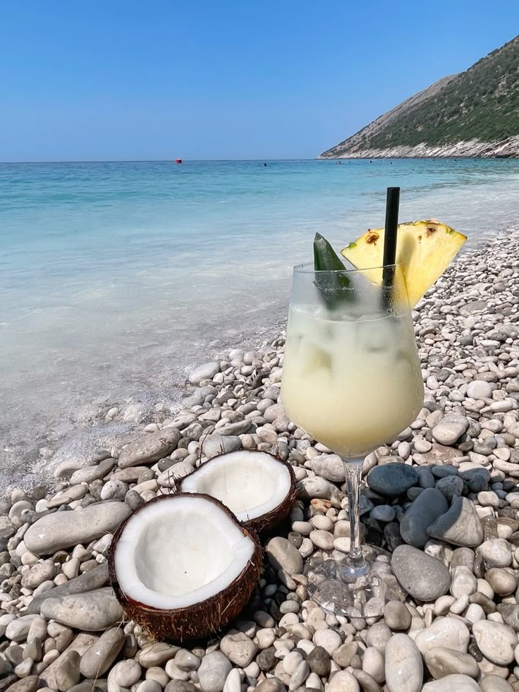 Chilled cocktail with pineapple garnish on a rocky beach beside coconut halves, ocean in the background.