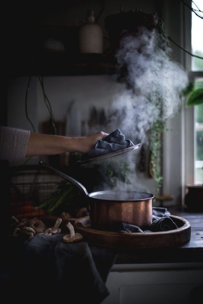 Steaming copper pot on stove with hand lifting lid, surrounded by mushrooms and rustic kitchen scene.