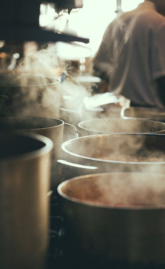 Steaming pots in a busy commercial kitchen with a chef preparing food in the background.