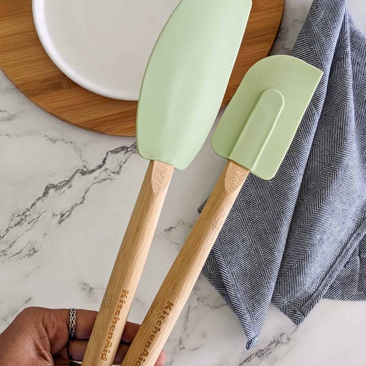 Green silicone spatulas with wooden handles on a marble countertop by a blue napkin and wooden board.