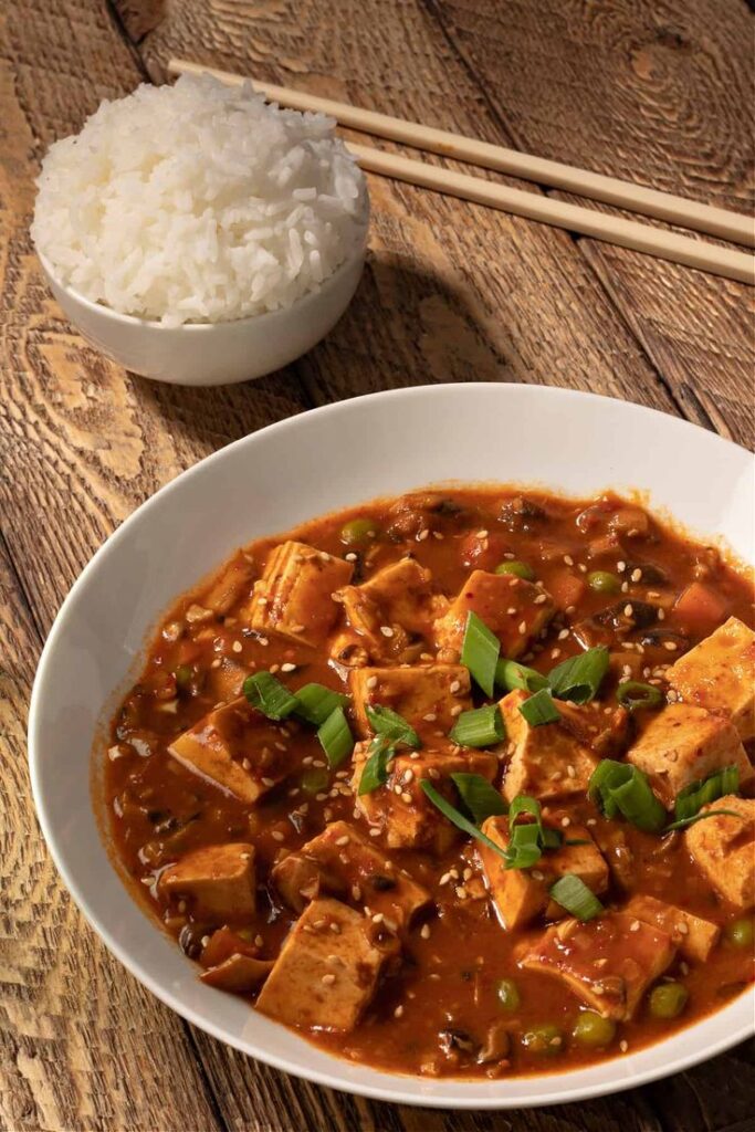 Spicy tofu stew with green onions and sesame seeds served alongside a bowl of steamed rice on a rustic table.