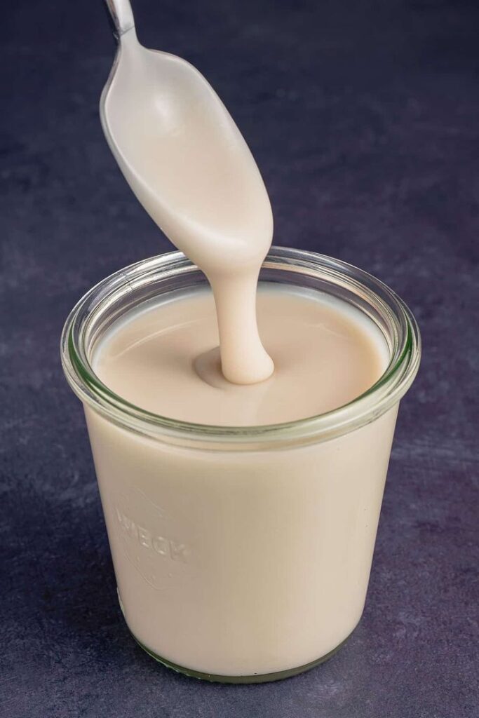 Creamy tahini being poured from a spoon into a glass jar on a dark surface.