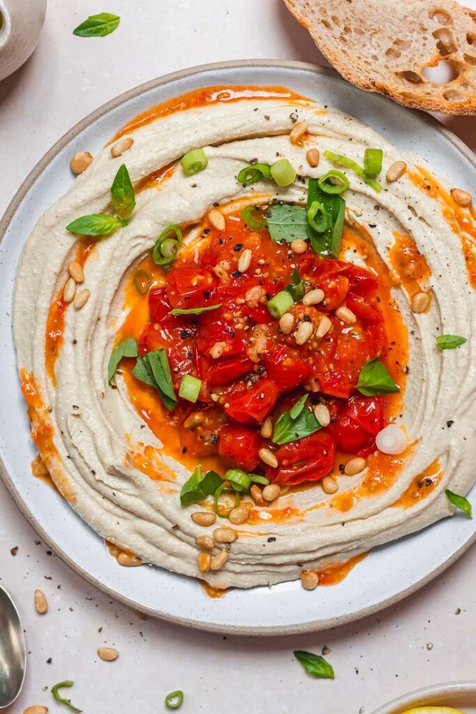 Creamy hummus topped with roasted tomatoes, pine nuts, and fresh herbs on a white plate.