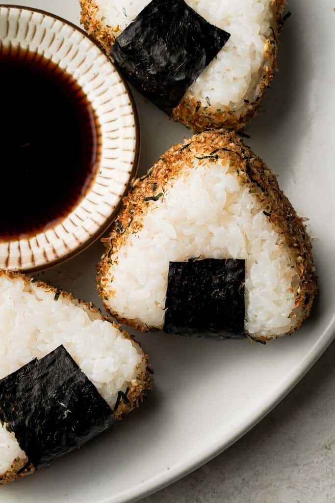 Japanese onigiri rice balls, wrapped in seaweed, served with soy sauce on a white plate.