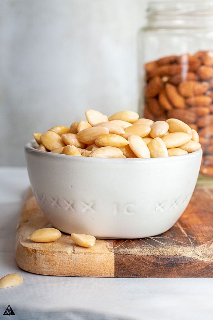 Bowl of blanched almonds on wooden board, with jar of unpeeled almonds in background, ready for snacking.