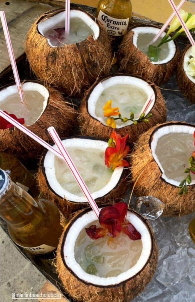 Coconut cocktails with colorful straws and flowers, surrounded by icy Coronas, offer a tropical beach vibe.