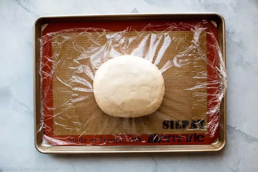 Dough ball wrapped in plastic on a baking sheet, ready for rising.