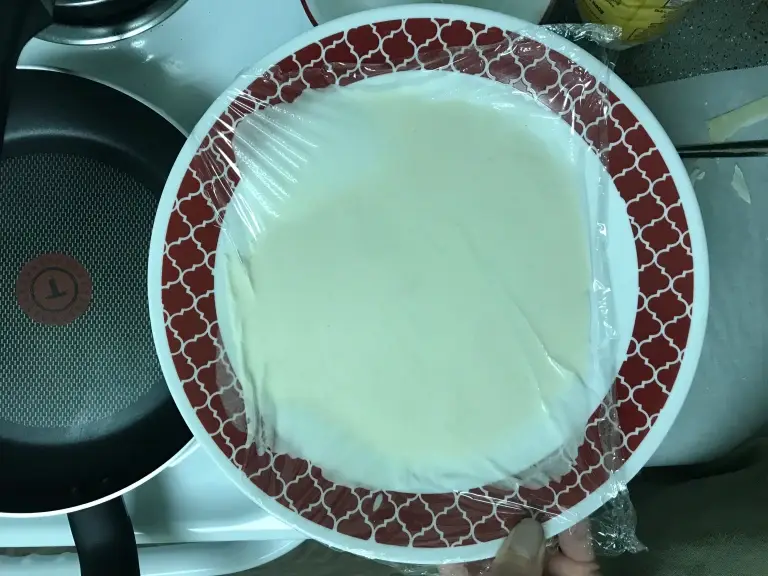 Batter in a decorative plate covered with plastic wrap, next to a non-stick frying pan on a stove.