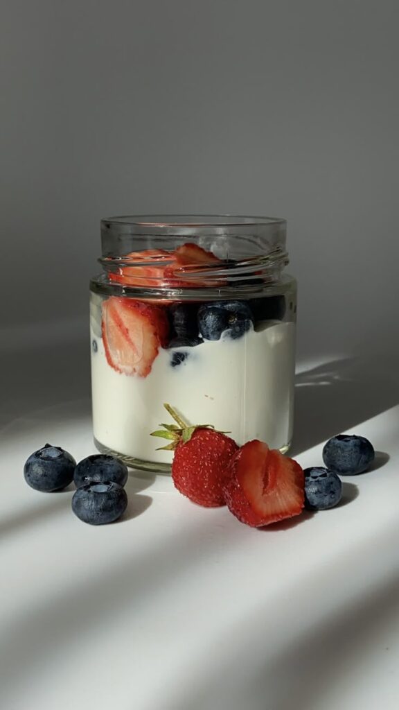 Jar of creamy yogurt topped with fresh strawberries and blueberries on a bright surface.