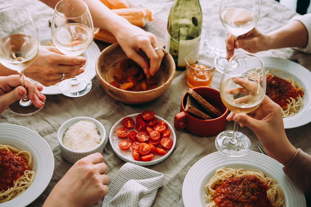 Friends enjoying a pasta meal with wine, featuring tomatoes, cheese, and bread on a cozy dining table.
