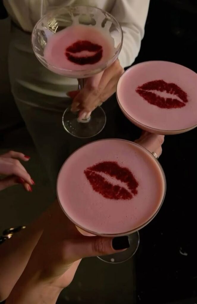 Pink cocktails with red lip print designs on foam, held by hands with manicured nails in a dim setting.
