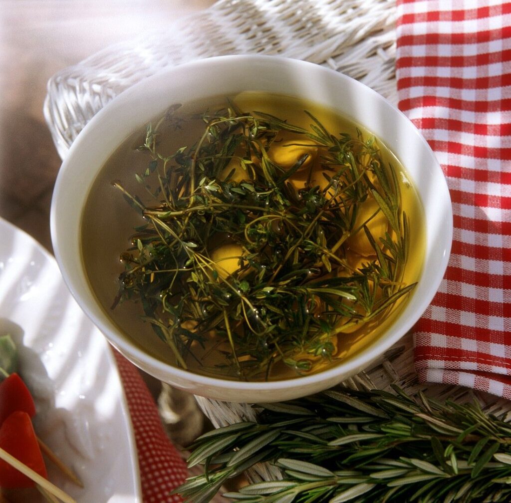 Bowl of seasoned olive oil with fresh herbs, rosemary, and garlic, accompanied by a red checkered napkin.