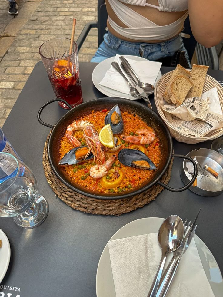 Seafood paella with mussels, shrimp, and lemon slices served on an outdoor table, accompanied by bread and a drink.