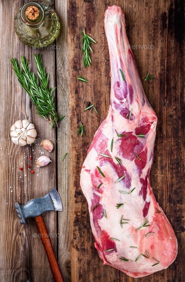 Raw leg of lamb with rosemary on a rustic board, surrounded by garlic, herbs, and a butcher's axe for cooking prep.
