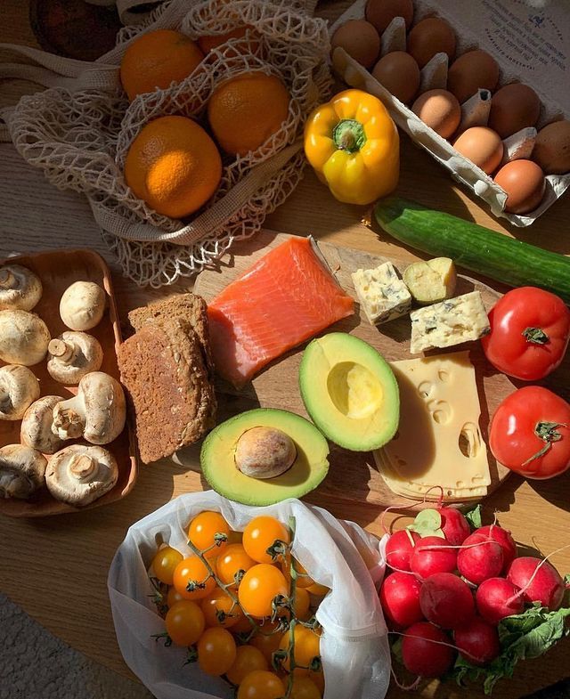 Assorted fresh produce and foods, including salmon, avocado, bread, cheese, veggies, and eggs, displayed on a wooden table.