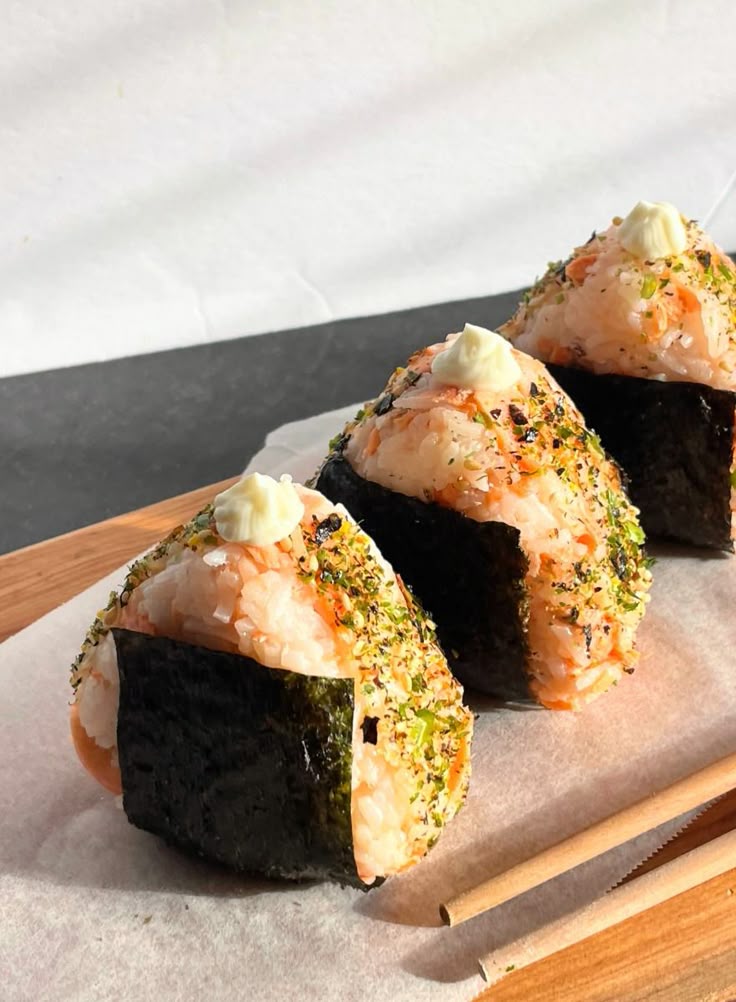 Three freshly made onigiri rice balls topped with seasoning and sauce, served on a wooden board with chopsticks.