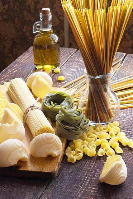 Assorted pasta types and olive oil on a rustic table, featuring spaghetti, shells, tagliatelle, and oil bottle.