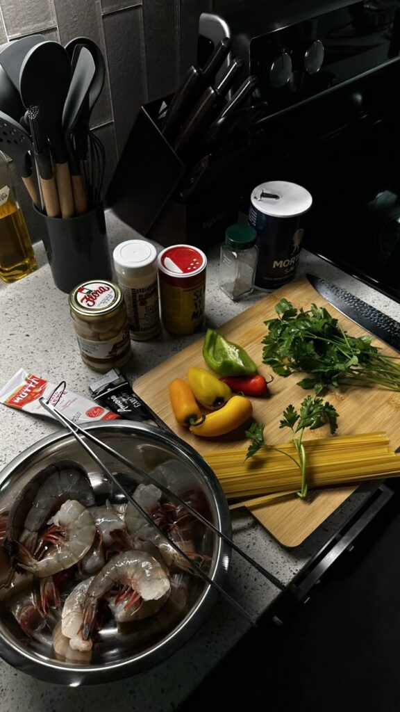Fresh ingredients for pasta: shrimp, bell peppers, parsley, and spices on a kitchen counter with utensils.