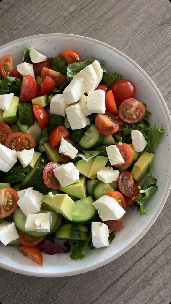 Fresh salad with cherry tomatoes, avocado, cucumber, mozzarella, and greens in a white bowl on wood background.