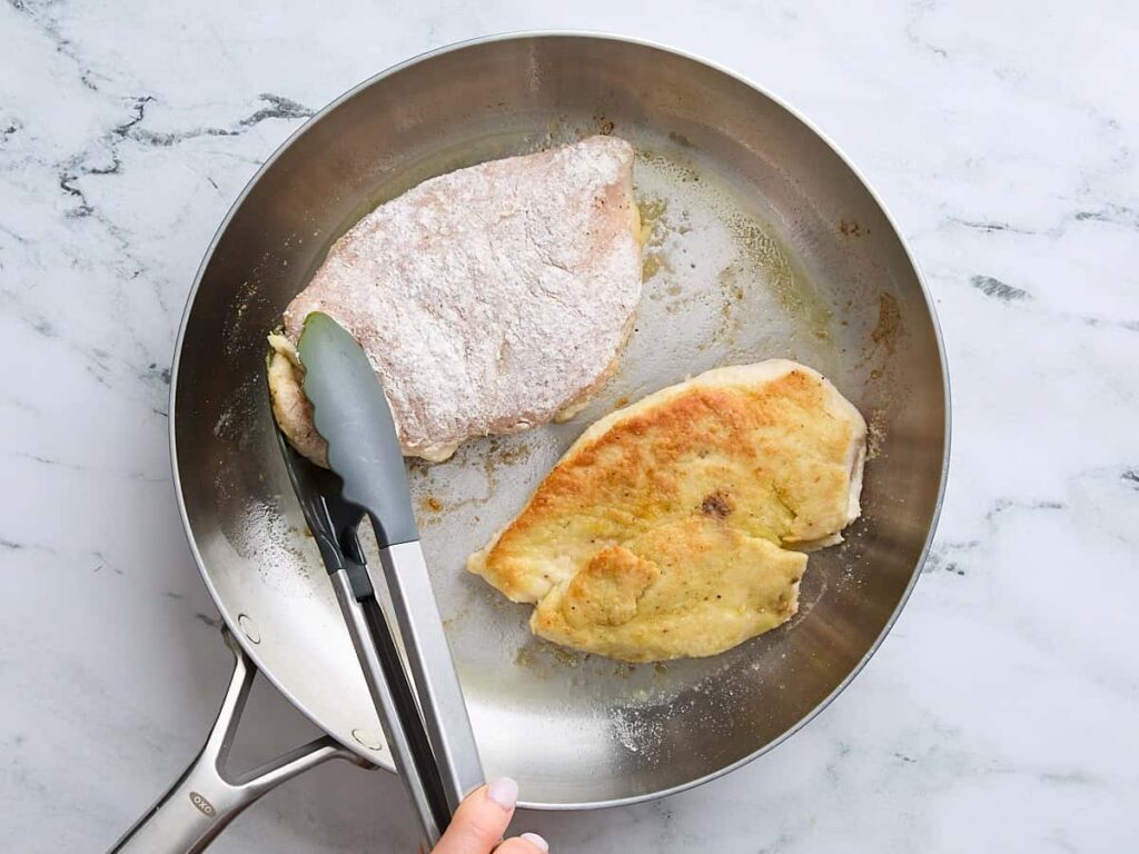 Cooking breaded chicken breasts in a stainless steel pan with tongs on a marble countertop.