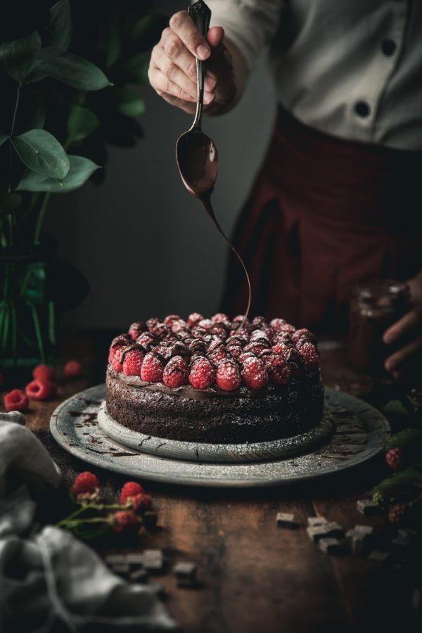 Hand drizzling chocolate over raspberry-topped cake on a rustic table, garnished with fresh raspberries and powdered sugar.