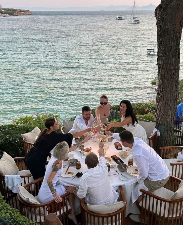 Group of friends toasting at a seaside restaurant with ocean view and yachts in the background.