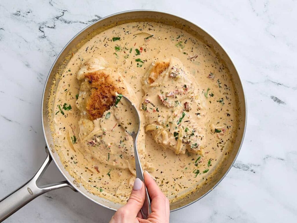 Creamy chicken in a skillet with a rich, herb-infused sauce being served with a spoon.