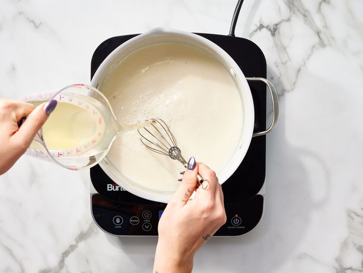Pouring liquid into a pot with a whisk on electric stovetop, cooking creamy sauce on marble countertop.