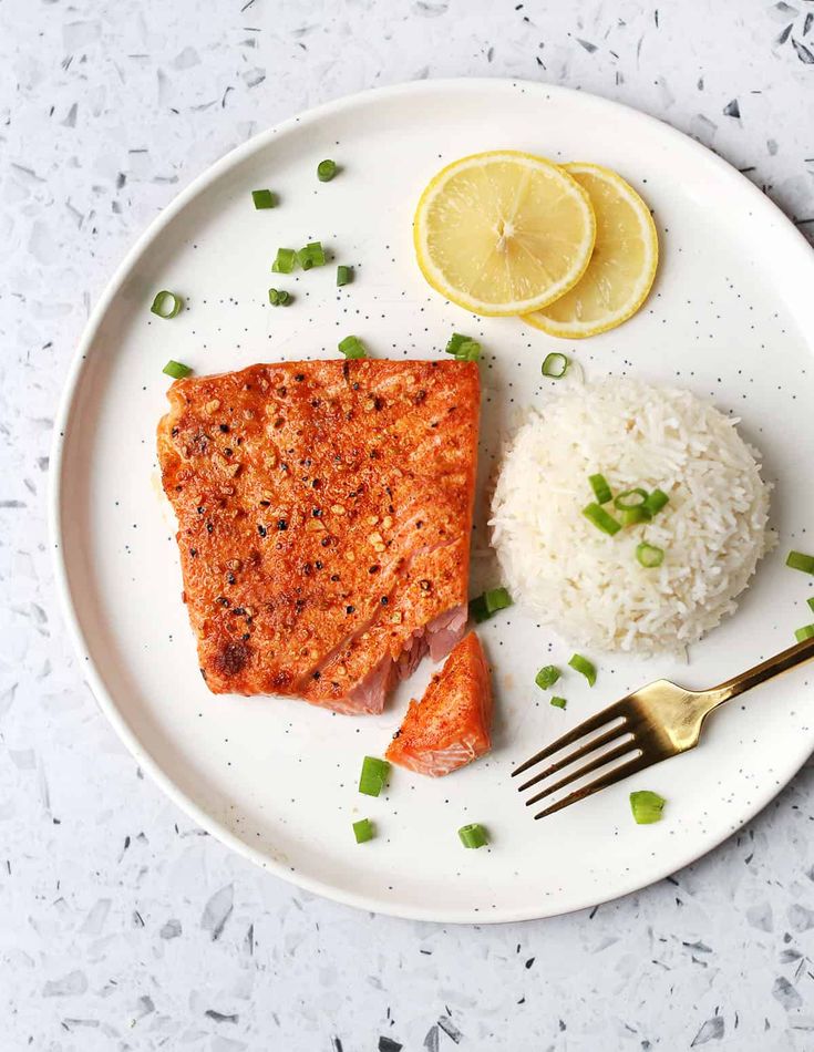 Grilled salmon with rice, lemon slices, and green onions on a white plate with a gold fork on a speckled surface.